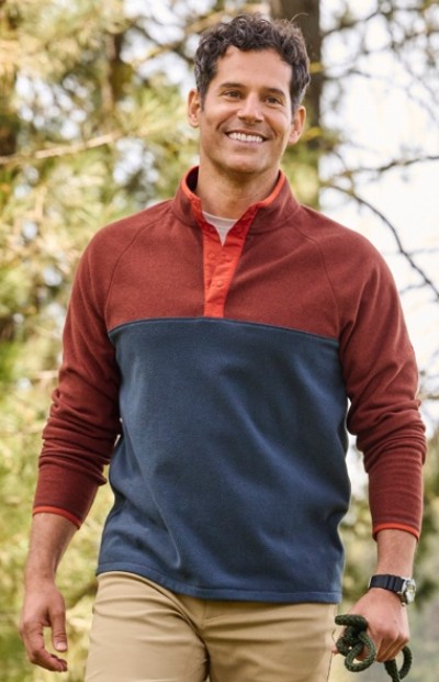 A man in red and blue color block fleece and beige pants holds a dog leash outdoors with trees in the background.