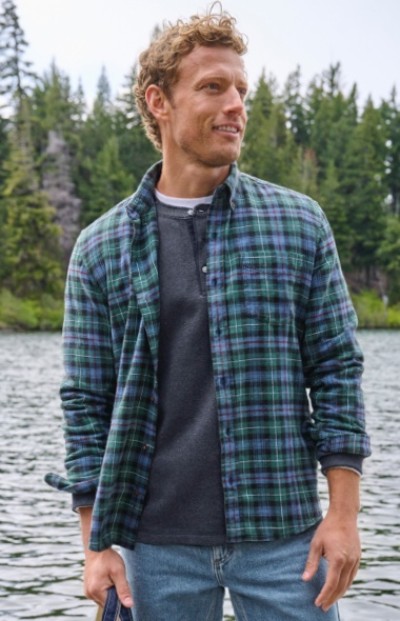 A man wearing a green and blue plaid shirt over a henley shirt stands by a lake with trees in the background.