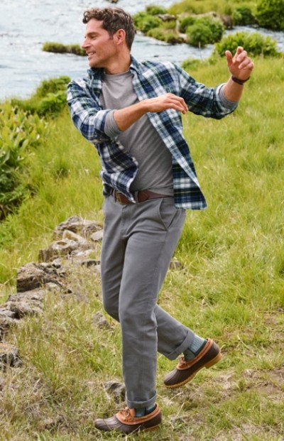 A man in blue and white plaid shirt and gray pants stands on grass near water with arms raised and one foot lifted.