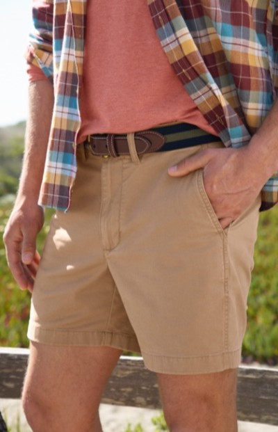 Cropped image of a man wearing a plaid shirt, pink t-shirt, and beige shorts with brown belt stands outdoors with one hand in pocket.