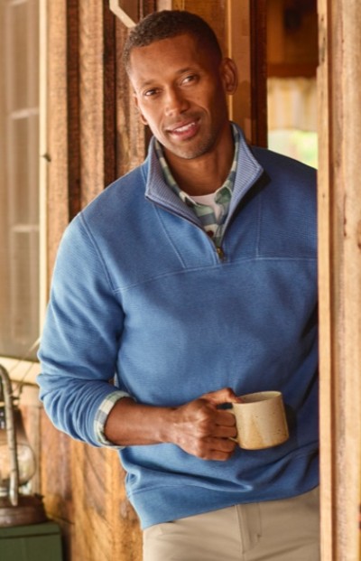 A man wearing a blue sweater holds a coffee mug while standing in an exterior doorway.