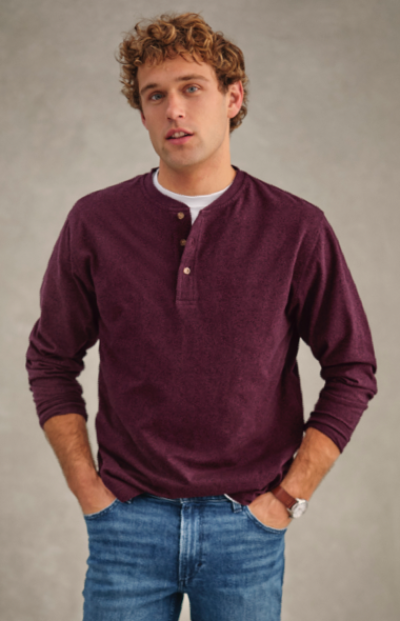 A man wearing a maroon long-sleeve henley shirt and blue jeans stands with hands in pockets against a plain background.