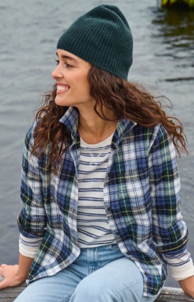 A woman with long curly hair in a green beanie, plaid shirt, striped tee, and jeans sits by the water.