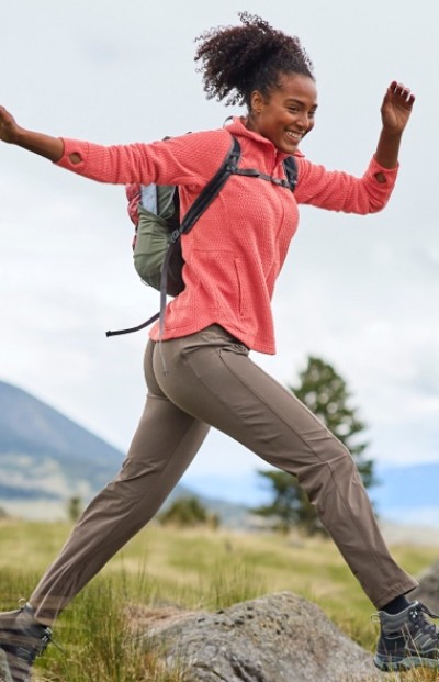 A woman in a pink top, tan pants and backpack hikes over rocks in a grassy area, with mountains and trees in the background.