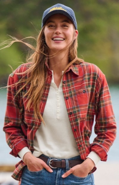 Woman wearing a red plaid shirt, white top, blue jeans, and corduroy baseball cap stands outdoors with hands in pockets.