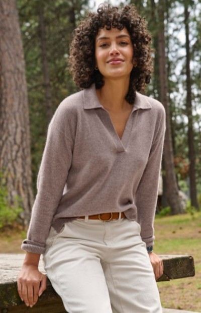 A woman in light brown sweater and white pants sits on a wooden bench outdoors, surrounded by trees.
