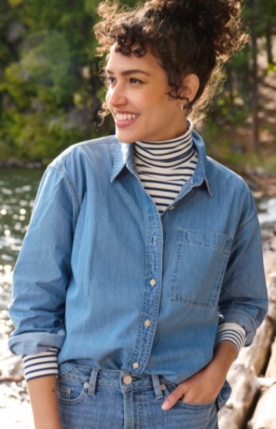 A woman with curly hair wearing a blue denim shirt over striped turtleneck stands outdoors near water and trees. 