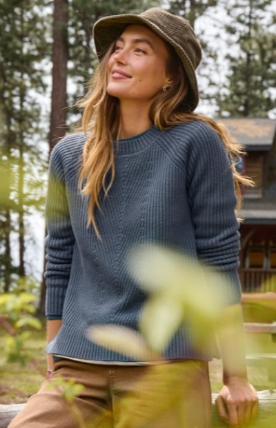 A woman wearing a blue sweater, brown pants and a wide brimmed hat stands outdoors near trees and a house.