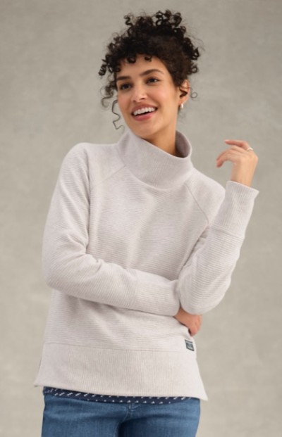 A woman wearing a light gray turtleneck and blue jeans stands against a plain background with one arm crossed.