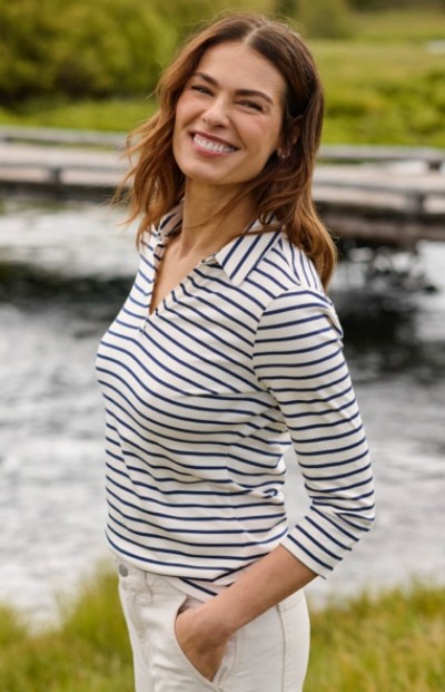 A woman in white and navy striped shirt and white pants stands near water, surrounded by greenery in an outdoor setting.