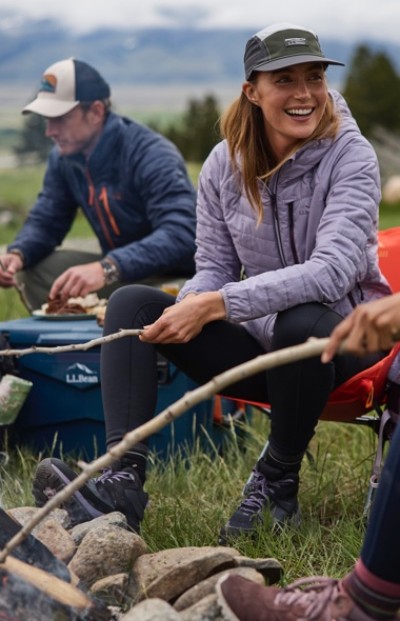Two people sit by a campfire roasting food on sticks, dressed in outdoor gear and surrounded by camping equipment.