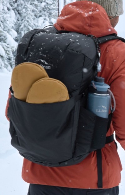 Person in red jacket and beige beanie carries an L.L.Bean backpack with snowshoes and a blue bottle in a snowy forest.