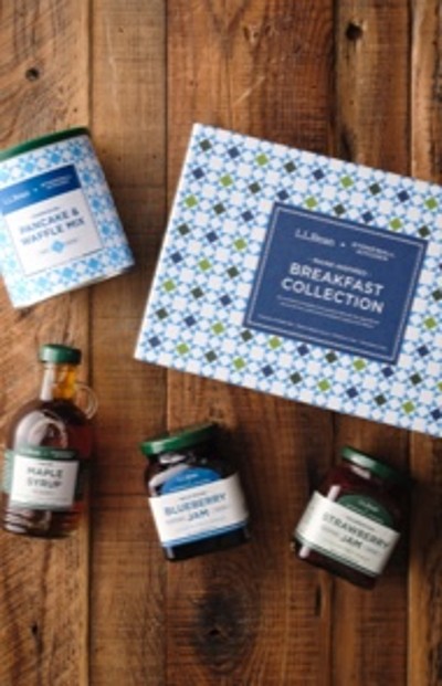 L L Bean breakfast collection with with three fruit preserve jars, maple syrup, and pancake & waffle mix on a wooden surface.