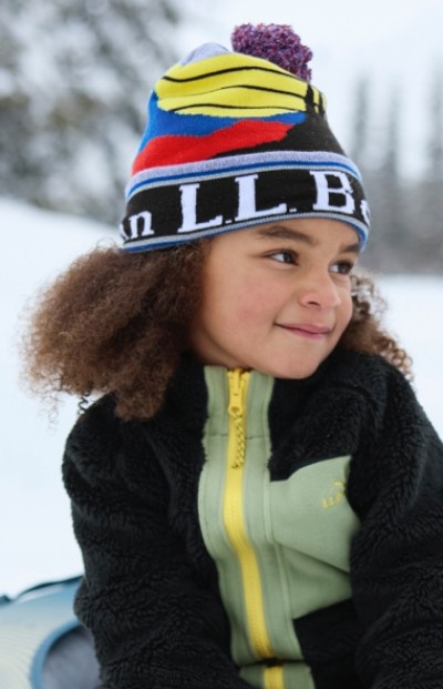 Child wearing a colorful beanie and black jacket with yellow zipper stands outdoors in a snowy setting.