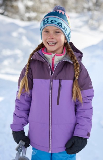 Smiling child in purple color block jacket, blue pants, beanie and gloves stands in the snow holding a helmet.