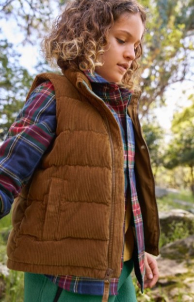 A child in brown corduroy vest and plaid shirt stands outdoors among trees and rocks in a natural setting.