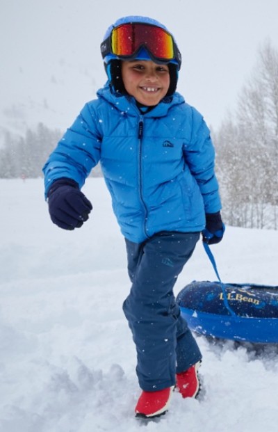 Child in blue winter gear, red boots, and helmet with goggles pulls an L.L.Bean sled through snowy landscape.