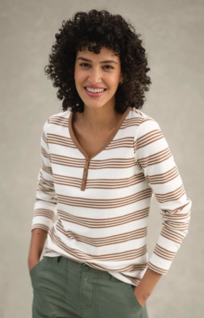 A woman with curly hair in a white and brown striped shirt and green pants stands with hands in pockets.