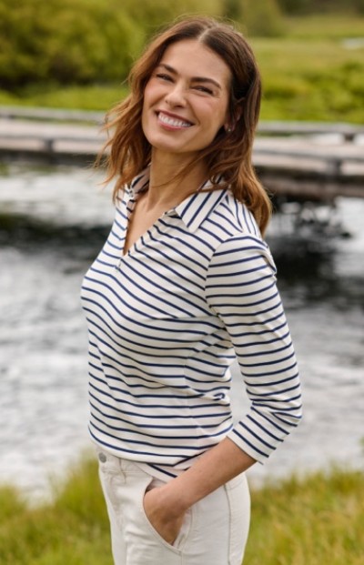 A woman wearing a white and navy striped shirt with white pants stands near water, surrounded by greenery.