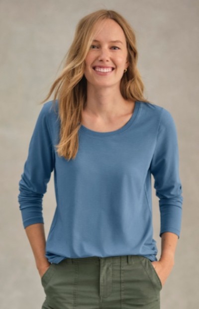 A woman in a blue long-sleeve shirt and green pants stands with hands in pockets.