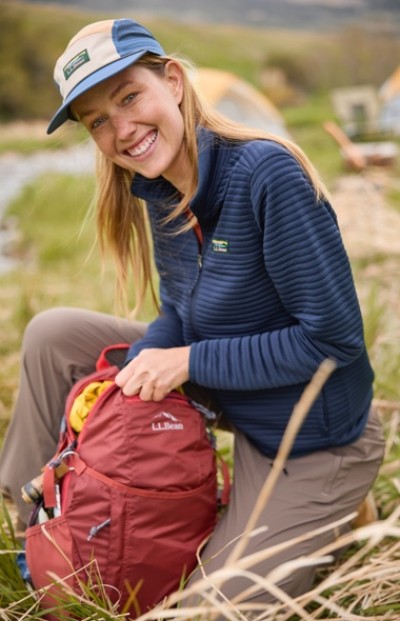 A person in blue jacket and cap kneels on grass with red L. L.Bean backpack; tent and camping gear visible in the background.