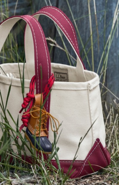 A mini red and white canvas tote bag with an L. L. Bean boot keychain attached to the handle sits in the grass.