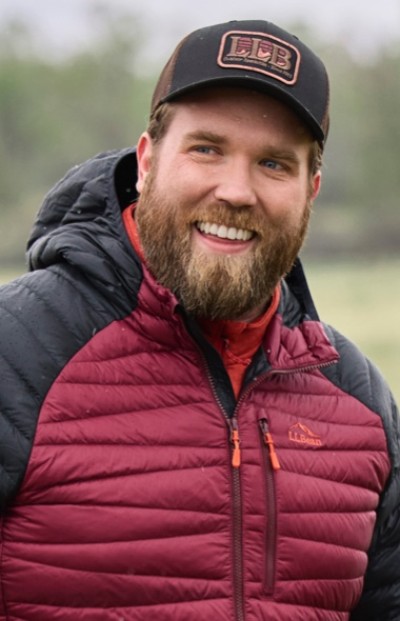 Person in red and black jacket and black cap stands outdoors with greenery in the background.
