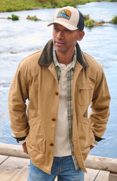 Person in tan jacket, plaid shirt, and cap stands on a wooden platform by a river, with greenery in the background.