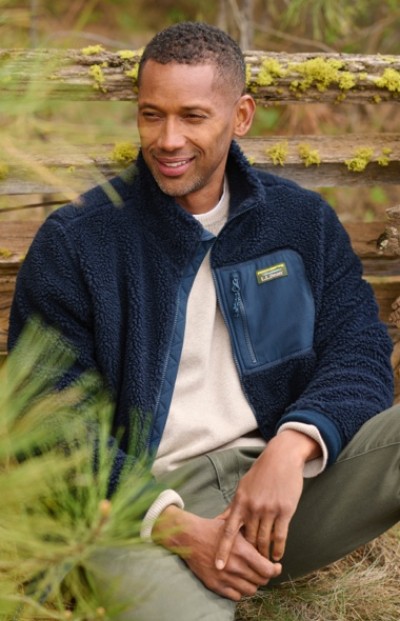 A man in navy fleece and light shirt sits outdoors on grass, with greenery and a wooden fence behind him.