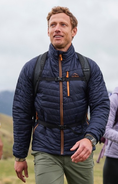 A man in blue quilted jacket and green pants hikes with a backpack under a cloudy sky, surrounded by grassy hills.