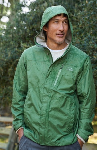 A person in green rain jacket with hood up stands outdoors with hands in pockets.
