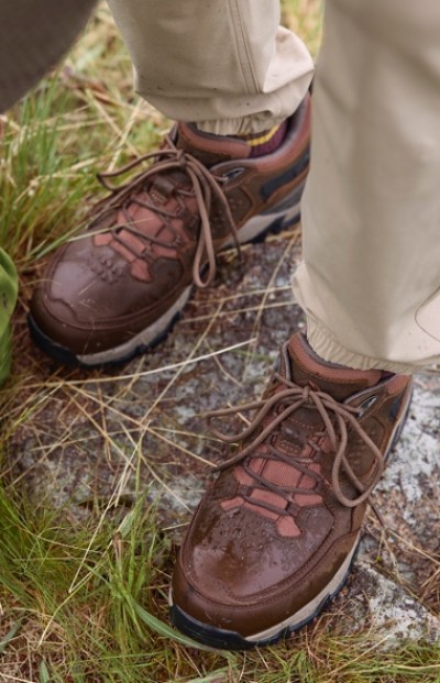Brown hiking shoes with laces worn by someone in beige pants, standing on rocky ground surrounded by grass.
