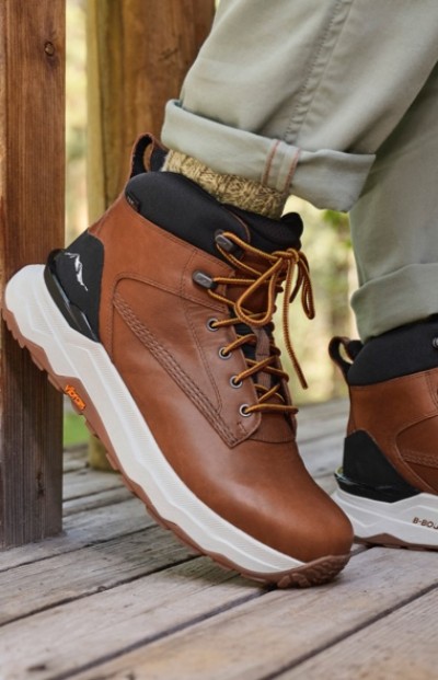 Close up of a person wearing brown hiking boots with white soles and black accents standing on a wooden deck.