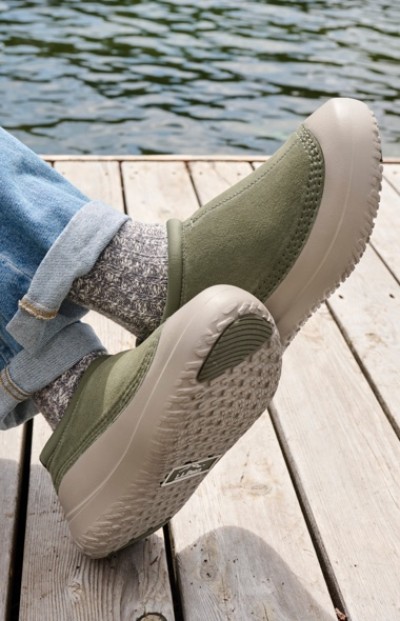 Close up of crossed feet wearing green slip-on shoes with gray socks resting on a wooden dock by the water.