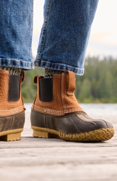 Close up of a person in brown L. L. Bean Chelsea boots and blue jeans stands on a wooden dock near water.