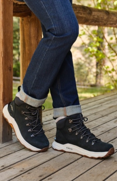 Person in rolled-up blue jeans and black hiking boots with white soles leans on a wooden deck.