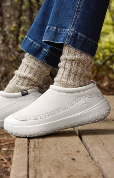 Close up of a person in white slip-on shoes, beige knitted socks, and blue jeans standing on a wooden surface outdoors.