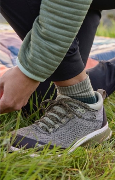 Close up of a person in gray sneakers and green jacket kneeling on grass and adjusting laces.