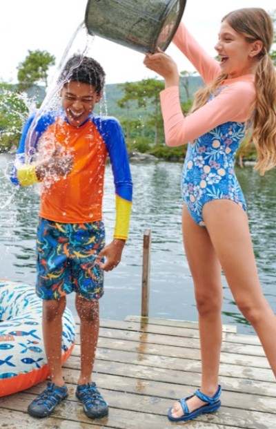 Child in blue and orange swim gear gets splashed with a bucket of water by another in floral swimsuit on a wooden dock.