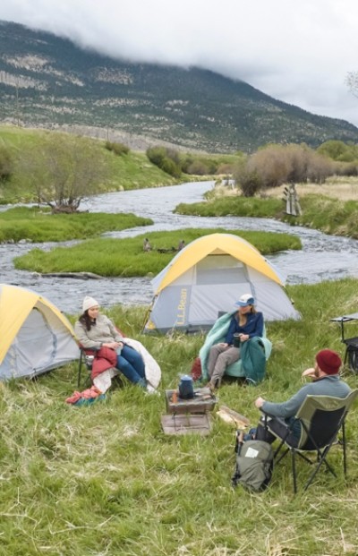Four people sit around a campfire near two tents by a river in a grassy area, with mountains in the background.