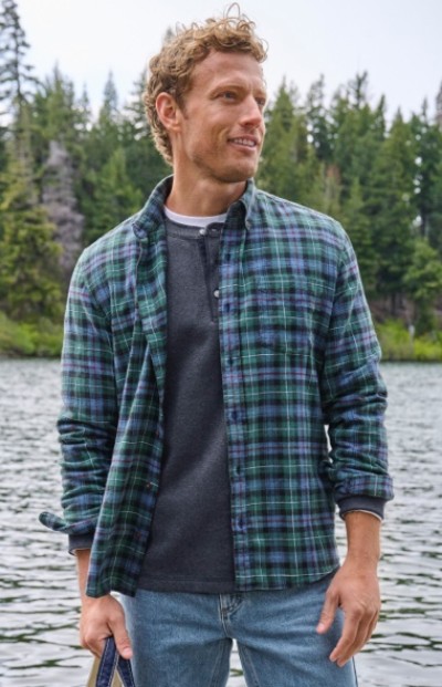 Man in plaid shirt stands by a lake with trees in the background, smiling and looking to the side.