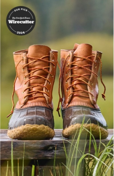 Brown leather L. L. Bean boots sit on a wooden surface, surrounded by green grass and a Wirecutter top pick badge.