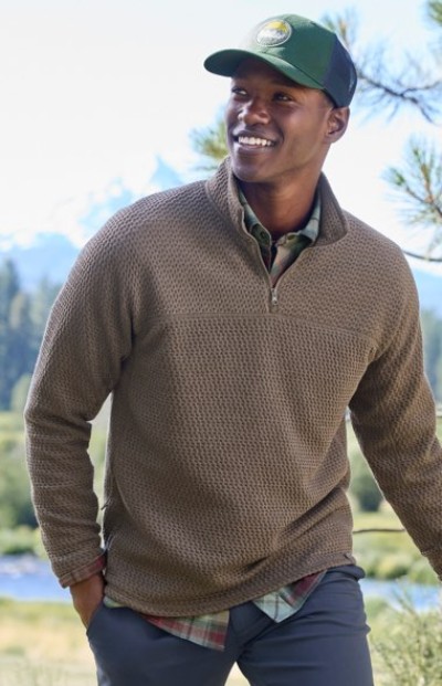 A person in a brown pullover, green cap, and plaid shirt stands outdoors with trees and mountains in the background.