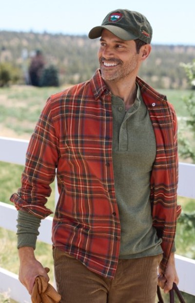 A person in a red plaid shirt and green cap holds a bag outdoors, with a white fence and greenery in the background.