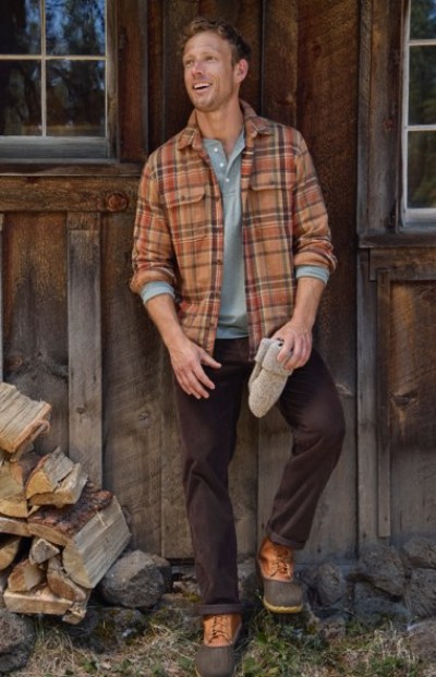 A person in a plaid shirt and brown pants stands by a wooden cabin, holding a hat, with stacked firewood nearby.