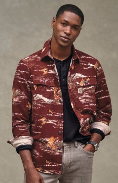 A person in a patterned maroon jacket and black shirt stands against a neutral background.