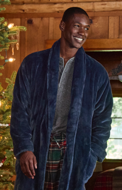 A person in a blue robe and plaid pants stands in a cozy cabin near a decorated Christmas tree.