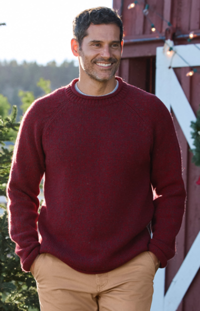 A person in a red sweater and beige pants stands near a barn with lights, surrounded by trees.