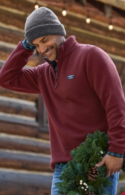 A person in a red sweater and gray beanie holds a holiday wreath in front of a log cabin decorated with string lights.