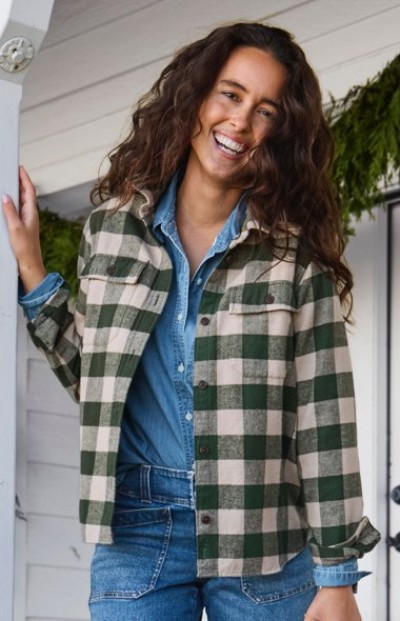 Person in green and beige checkered jacket over denim shirt stands in front of a white wall with greenery.